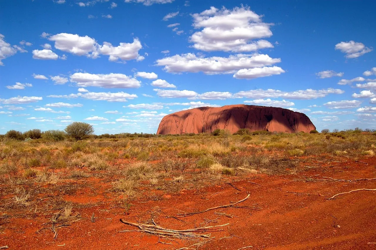 Der Uluru am Horizont, davor rote Erde und vereinzelte Büsche