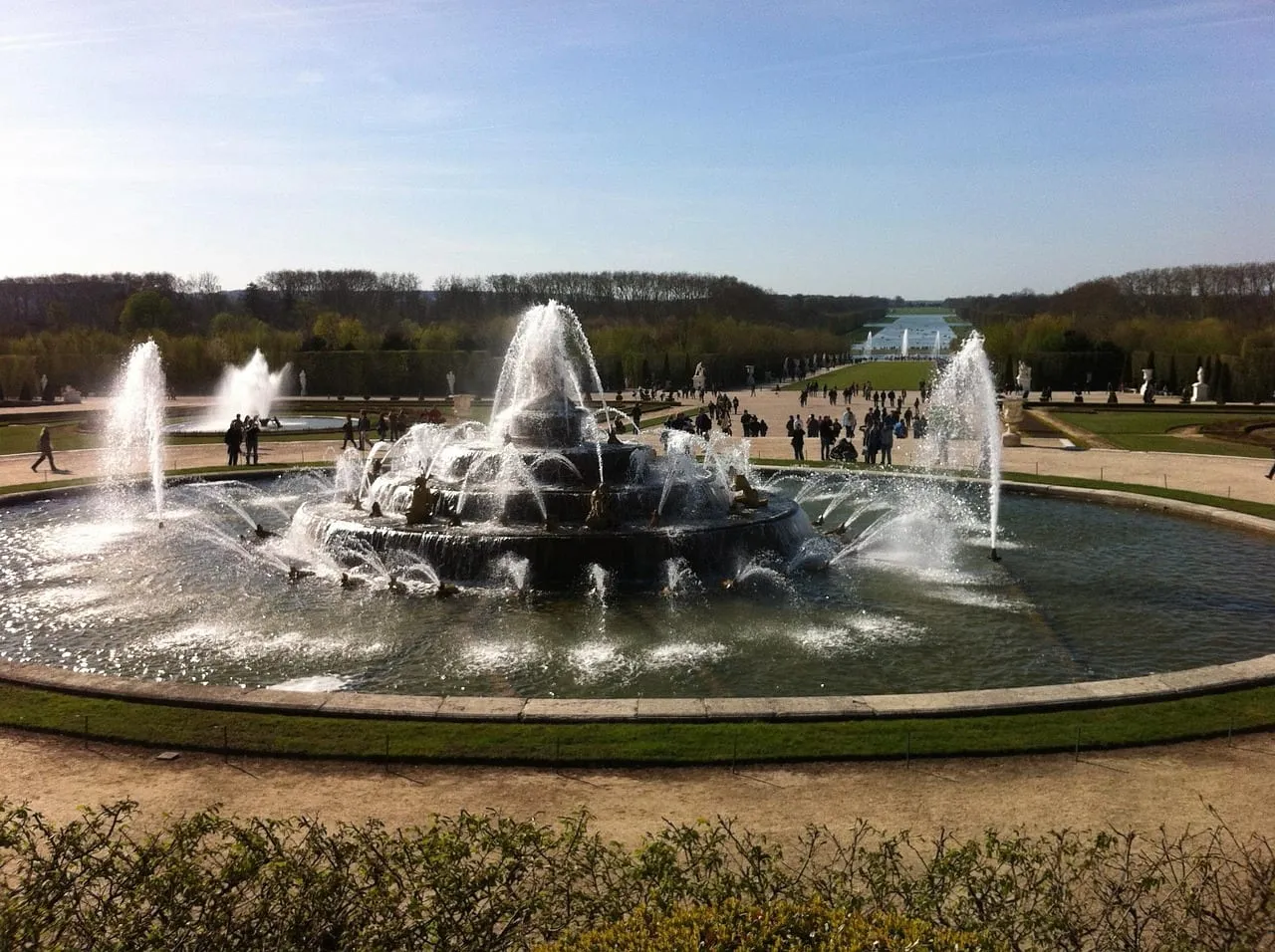 Ein Brunnen mit voll laufenden Wasserspielen in den Gärten, dahinter der weite Blick zum Kanal